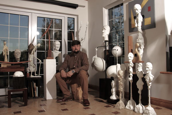 Valeriu Cazacevschi seated among his sculptures, with paintings on the wall.
