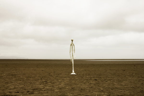 A pale, elongated sculpture photographed alone on an open beach beneath a cloudy sky.