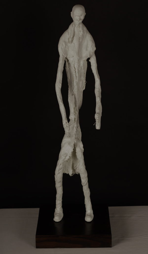 Elongated Sculpture by Valeriu Cazacevschi. A pale standing figure with very long arms and legs stands on a dark rectangular wooden base.