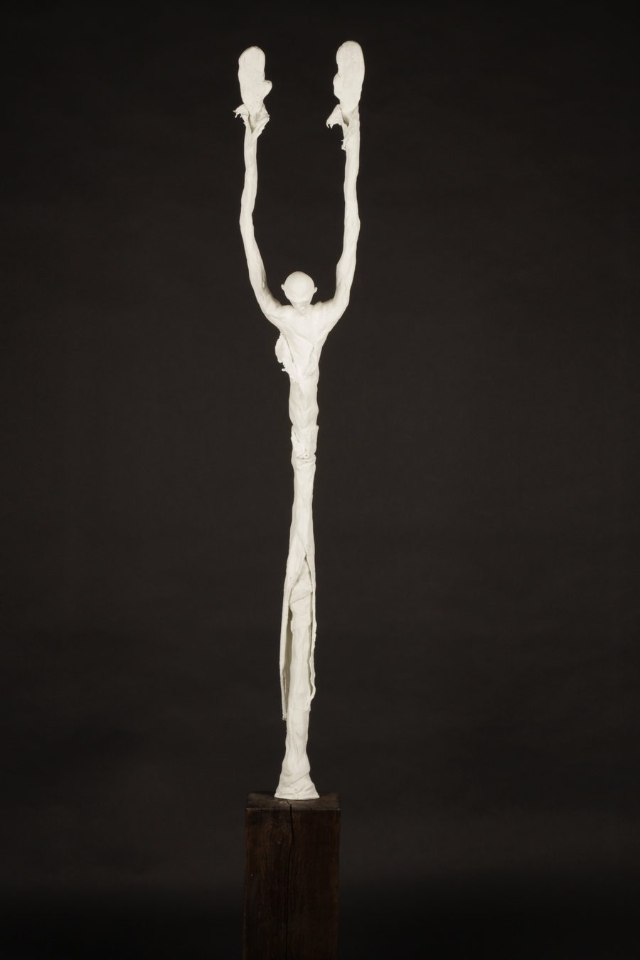 The Monk (2023) by Valeriu Cazacevschi. A tall pale figure raises both arms vertically above its head on a dark wooden support.